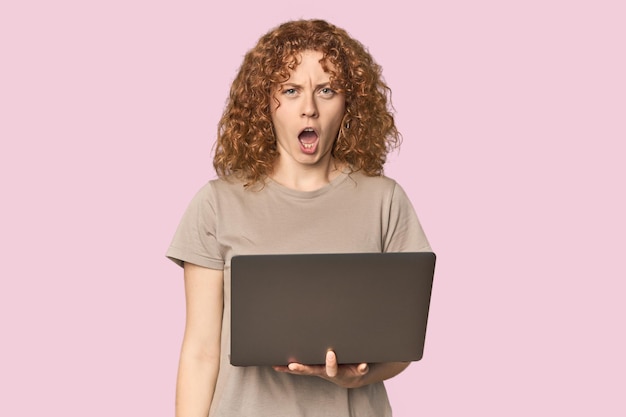 Joven mujer caucásica pelirroja con computadora portátil gritando muy enojada y agresiva