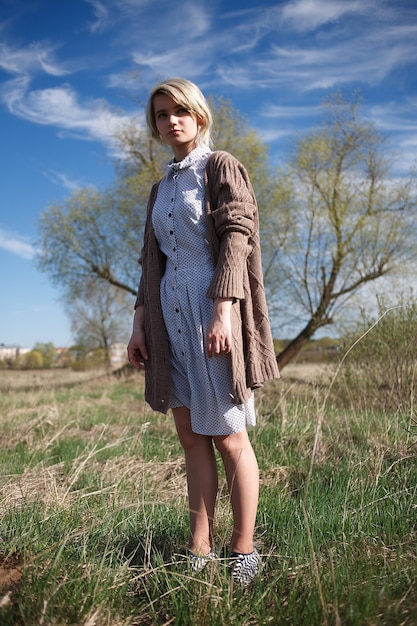 Joven mujer caucásica con pelo corto en vestido manchado y suéter de pie en el campo en un día soleado