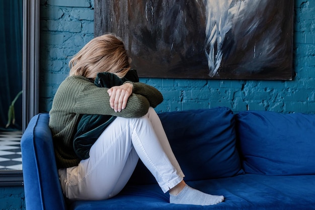 Foto joven mujer caucásica pensativa en suéter verde y jeans blancos sentado en el sofá con los ojos cerrados en la elegante sala de estar azul