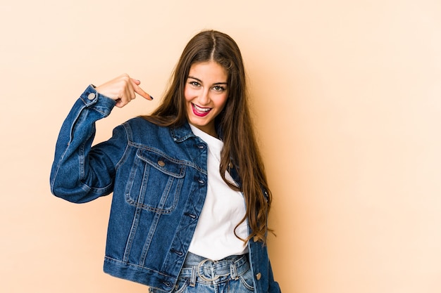 Joven mujer caucásica en persona beige apuntando con la mano a un espacio de copia de camisa, orgulloso y seguro