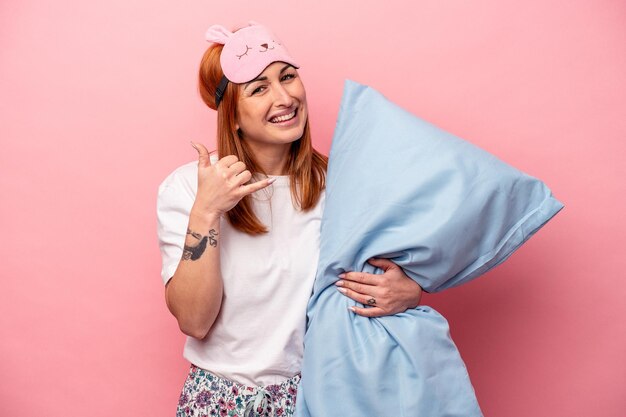 Joven mujer caucásica con un pijama sosteniendo una almohada aislada en un fondo rosa mostrando un gesto de llamada de teléfono móvil con los dedos