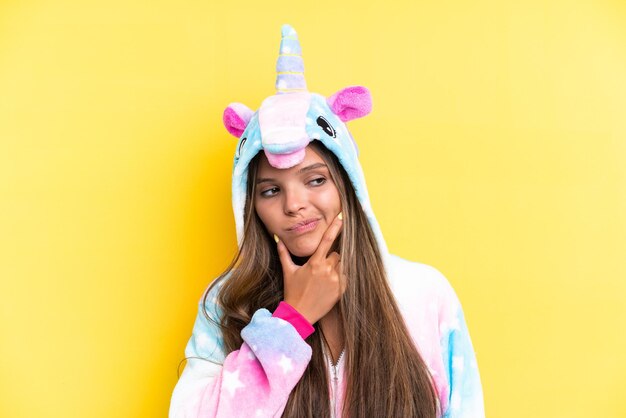 Joven mujer caucásica con un pijama de unicornio aislado de fondo amarillo que tiene dudas