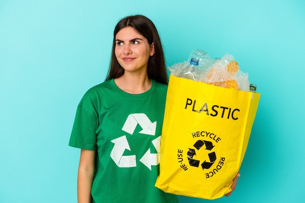 Joven mujer caucásica de plástico reciclado aislado en la pared azul soñando con lograr metas y propósitos