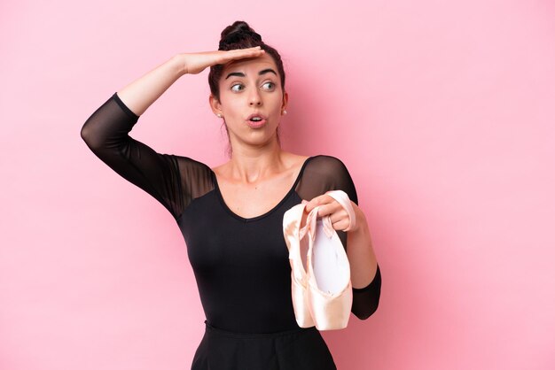 Joven mujer caucásica practicando ballet aislada de fondo rosa haciendo un gesto sorpresa mientras mira a un lado