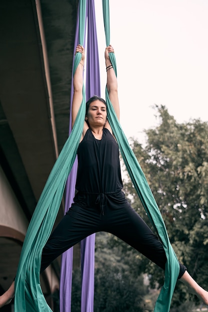 Foto joven mujer caucásica practicando rendimiento de sedas aéreas colgando de un puente en un parque de la ciudad, rodeado de naturaleza.