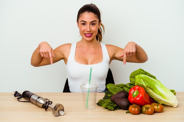 Joven mujer caucásica preparando un batido saludable con verduras apunta hacia abajo con los dedos, sentimiento positivo.