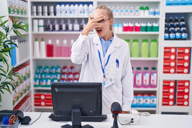 Joven mujer caucásica que trabaja en una farmacia mirando en shock cubriendo la cara y los ojos con la mano mirando a través de los dedos con expresión avergonzada