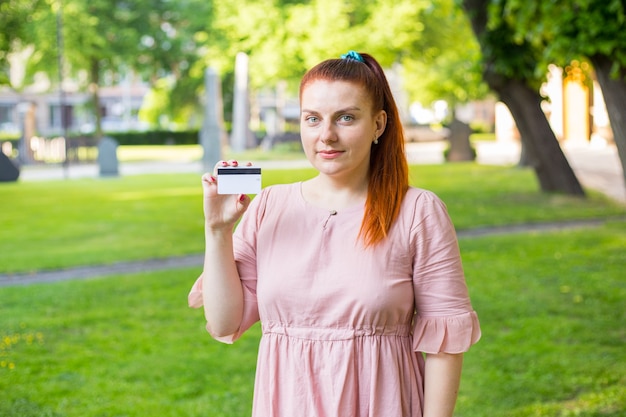 Joven mujer caucásica quedarse en el parque y mostrar una tarjeta de crédito blanca con una línea de imán negro.