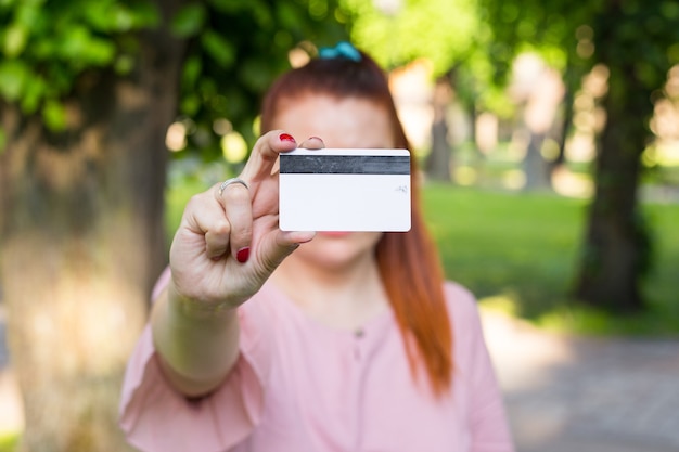 Joven mujer caucásica quedarse en el parque y mostrar una tarjeta de crédito blanca con una línea de imán negro.