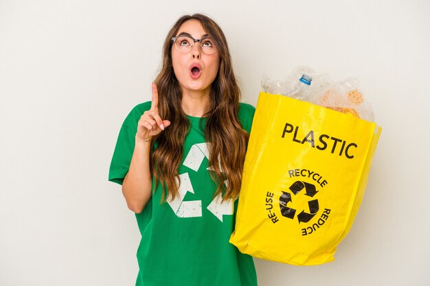 Joven mujer caucásica reciclando un lleno de plástico aislado sobre fondo blanco apuntando al revés con la boca abierta.