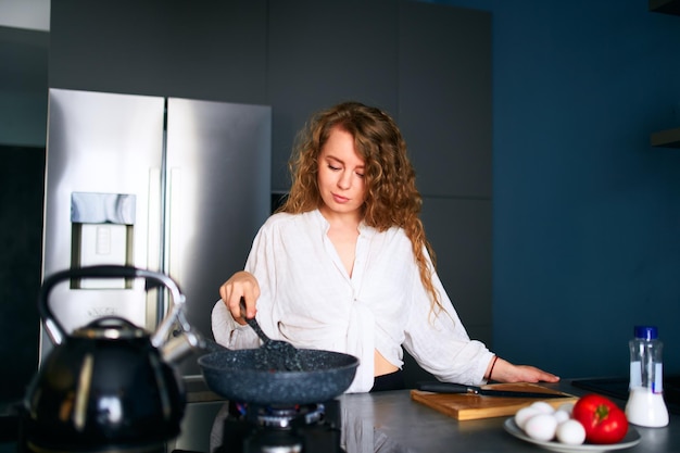 Joven mujer caucásica rizada sonriente cocina el desayuno en una sartén joven mujer adulta haciendo