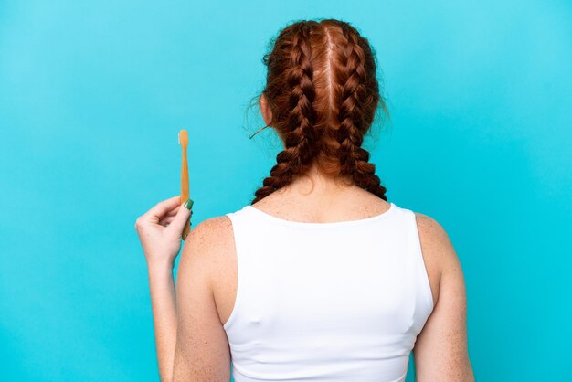 Foto joven mujer caucásica rojiza cepillarse los dientes aislado sobre fondo azul en posición trasera