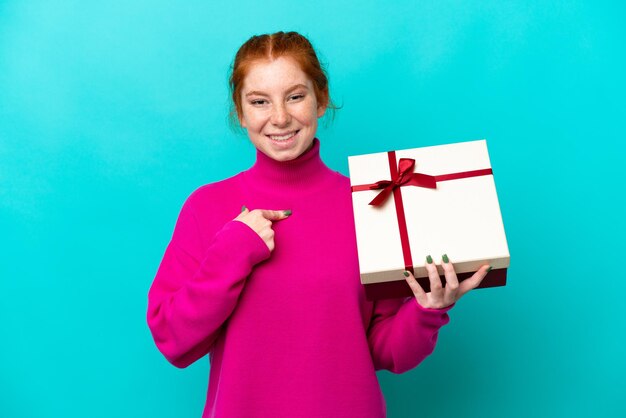 Joven mujer caucásica rojiza sosteniendo un regalo aislado de fondo azul con expresión facial sorpresa