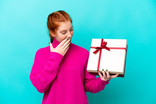 Joven mujer caucásica rojiza sosteniendo un regalo aislado de fondo azul con sorpresa y expresión facial conmocionada