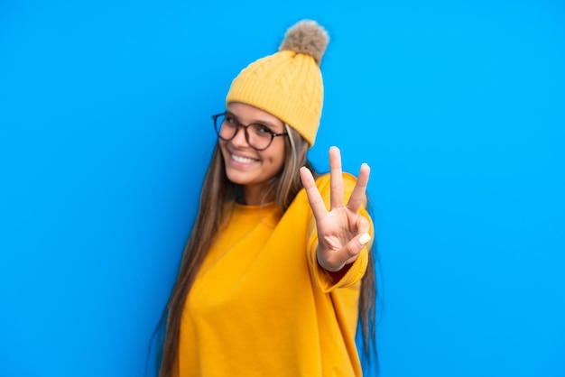 Joven mujer caucásica con ropa de invierno aislada de fondo azul feliz y contando tres con los dedos