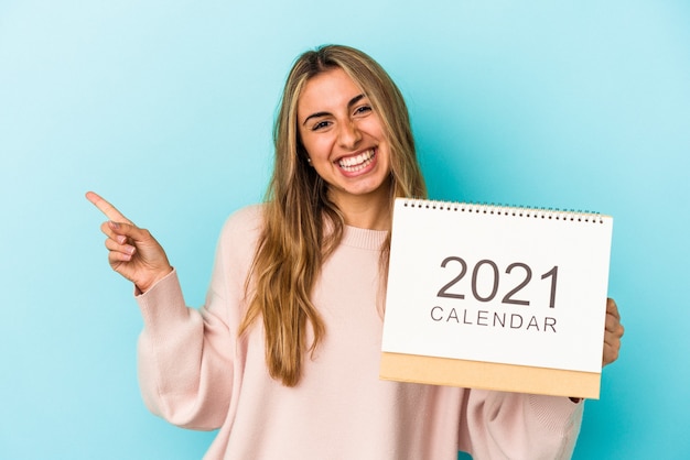 Joven mujer caucásica rubia agujereando un calendario aislado sonriendo y apuntando a un lado, mostrando algo en el espacio en blanco.