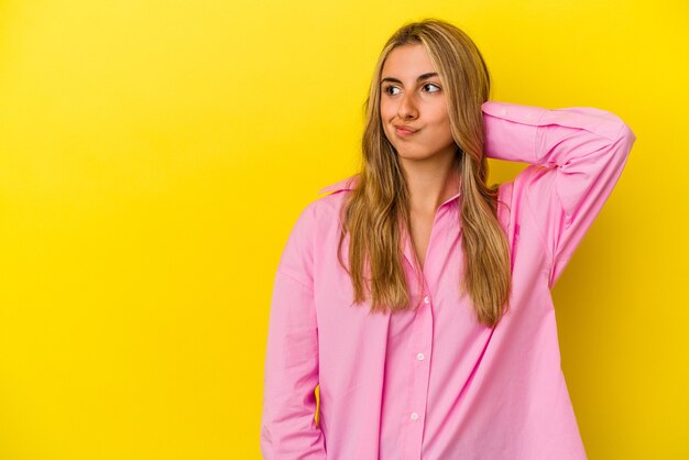 Joven mujer caucásica rubia aislada sobre fondo amarillo tocando la parte posterior de la cabeza, pensando y haciendo una elección.