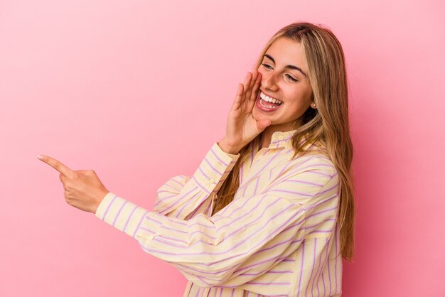 Joven mujer caucásica rubia aislada sobre fondo rosa diciendo un chisme, apuntando al lado informando algo.