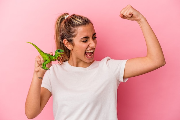 Joven mujer caucásica rubia sosteniendo una miniatura de dinosaurio aislada sobre fondo rosa levantando el puño después de una victoria, concepto ganador.