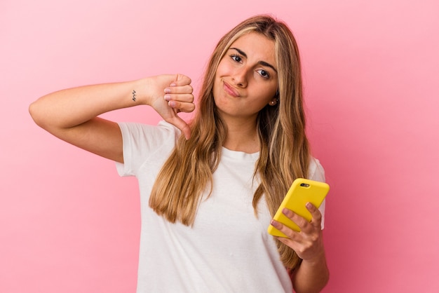 Joven mujer caucásica rubia sosteniendo un teléfono móvil amarillo aislado mostrando un gesto de aversión, pulgares hacia abajo