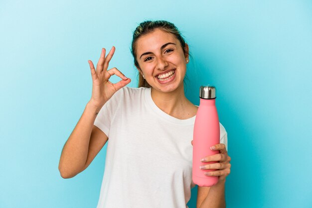 Foto joven mujer caucásica rubia sosteniendo un termo aislado sobre fondo azul alegre y confiado mostrando gesto ok.