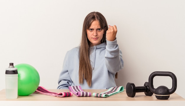 Joven mujer caucásica sentada en una mesa con equipamiento deportivo aislado sobre fondo blanco mostrando el puño a la cámara, expresión facial agresiva.