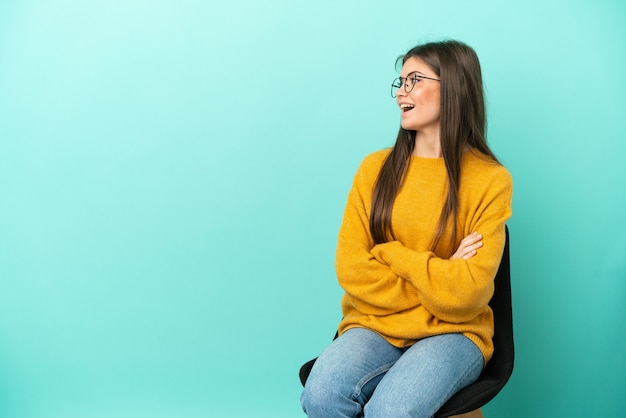 Joven mujer caucásica sentada en una silla aislada en la pared azul feliz y sonriente