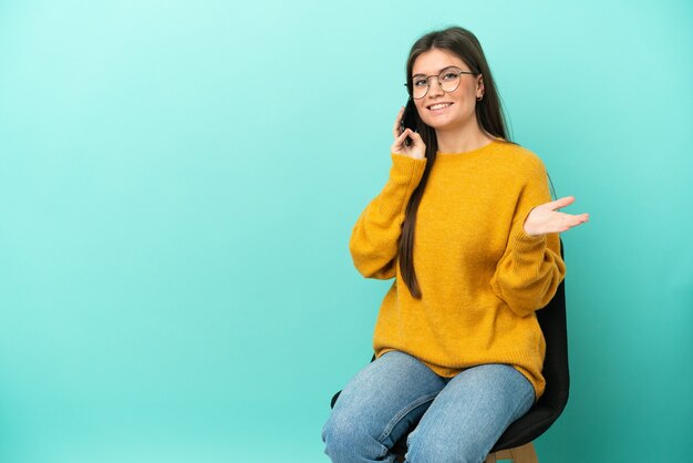 Joven mujer caucásica sentada en una silla aislada en la pared azul manteniendo una conversación con el teléfono móvil con alguien