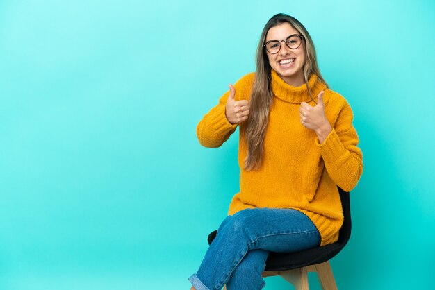 Joven mujer caucásica sentada en una silla aislada con los pulgares para arriba gesto y sonriendo