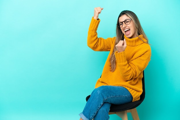 Joven mujer caucásica sentada en una silla aislada sobre fondo azul celebrando una victoria