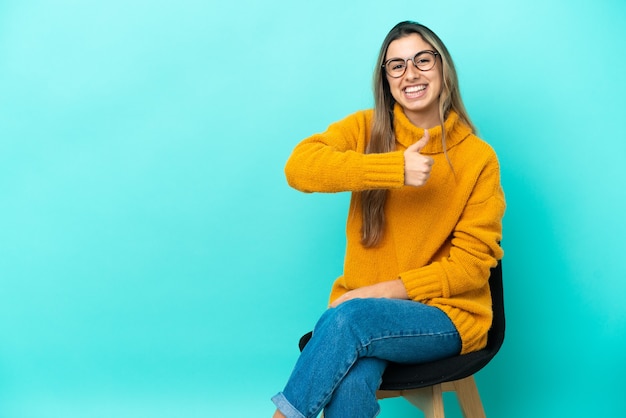 Joven mujer caucásica sentada en una silla aislada sobre fondo azul dando un pulgar hacia arriba gesto