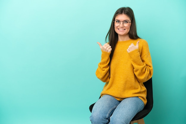 Joven mujer caucásica sentada en una silla aislada sobre fondo azul con gesto de pulgar hacia arriba y sonriendo