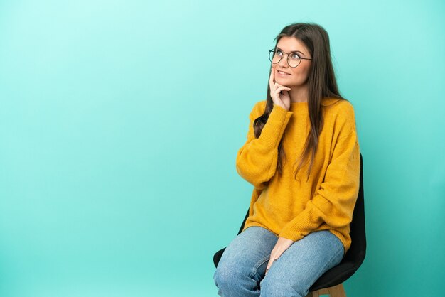 Joven mujer caucásica sentada en una silla aislada sobre fondo azul pensando en una idea mientras mira hacia arriba