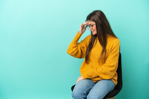 Joven mujer caucásica sentada en una silla aislada sobre fondo azul riendo