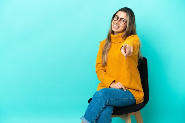 Joven mujer caucásica sentada en una silla aislada sobre fondo azul le señala con el dedo con una expresión de confianza