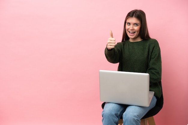 Joven mujer caucásica sentada en una silla con su computadora portátil aislada con los pulgares hacia arriba porque algo bueno ha sucedido