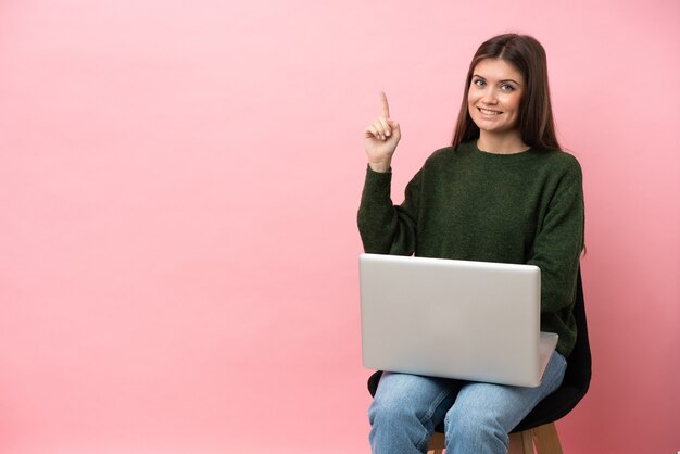 Joven mujer caucásica sentada en una silla con su computadora portátil aislada sobre fondo rosa mostrando y levantando un dedo en señal de lo mejor