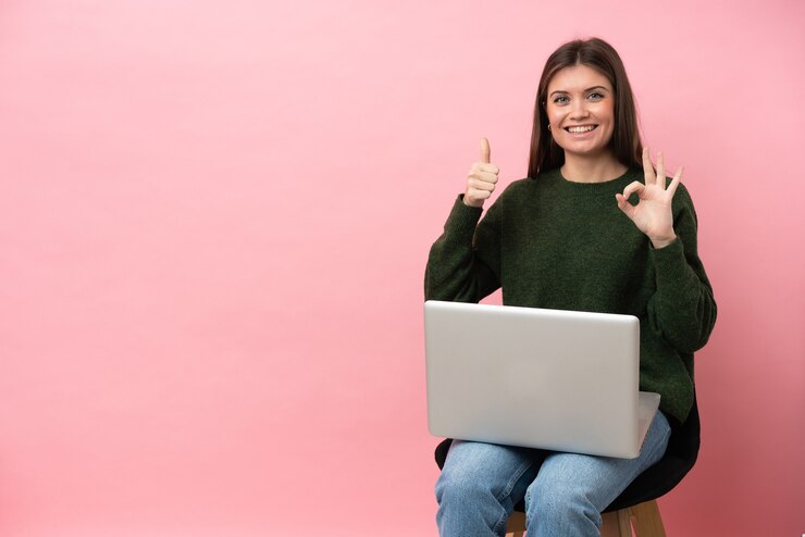 Foto joven mujer caucásica sentada en una silla con su computadora portátil aislada sobre fondo rosa mostrando el signo de ok y el pulgar hacia arriba gesto