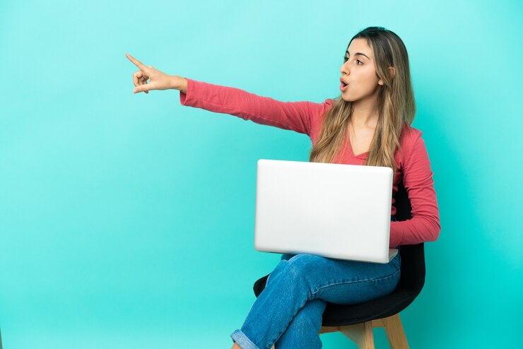 Foto joven mujer caucásica sentada en una silla con su pc aislado sobre fondo azul apuntando hacia afuera