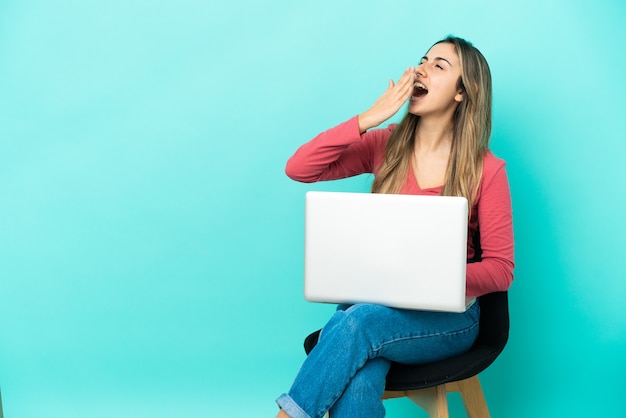 Joven mujer caucásica sentada en una silla con su pc aislado sobre fondo azul bostezando y cubriendo la boca abierta con la mano