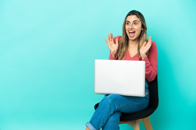 Joven mujer caucásica sentada en una silla con su pc aislado sobre fondo azul con expresión facial sorpresa