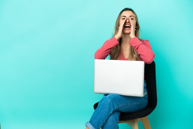 Joven mujer caucásica sentada en una silla con su pc aislado sobre fondo azul gritando y anunciando algo