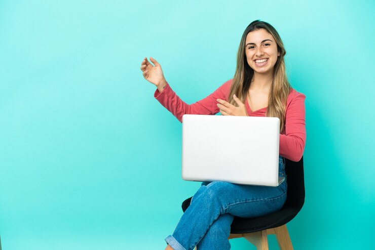 Foto joven mujer caucásica sentada en una silla con su pc aislado sobre fondo azul haciendo gesto de guitarra