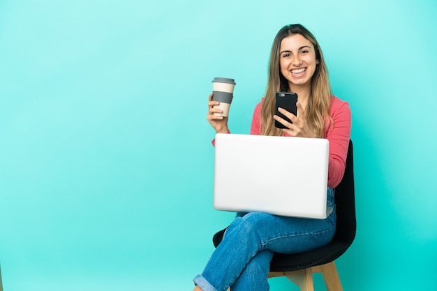 Joven mujer caucásica sentada en una silla con su pc aislado sobre fondo azul sosteniendo café para llevar y un móvil