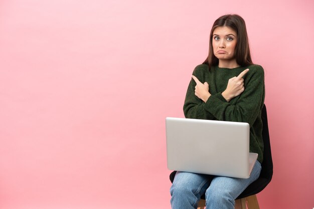 Joven mujer caucásica sentada en una silla con su portátil aislado apuntando a los laterales teniendo dudas