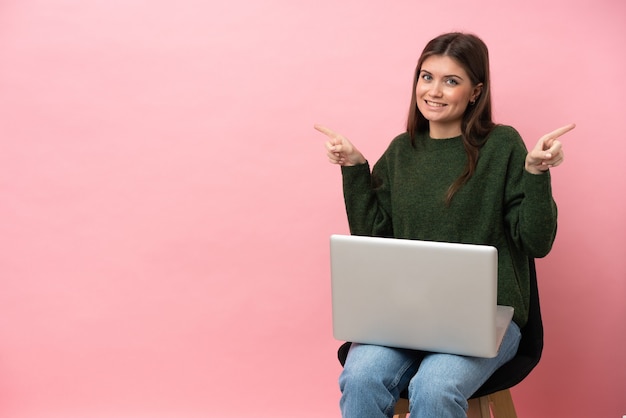 Joven mujer caucásica sentada en una silla con su portátil aislado sobre fondo rosa dedo acusador hacia los laterales y feliz