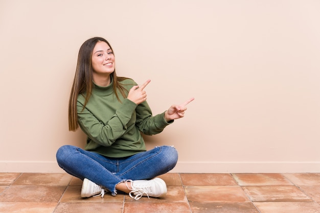 Joven mujer caucásica sentada en el suelo aislado emocionado apuntando con los dedos índices.