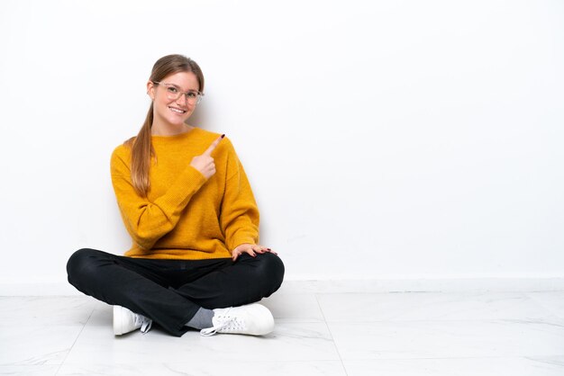 Joven mujer caucásica sentada en el suelo aislado sobre fondo blanco apuntando hacia el lado para presentar un producto