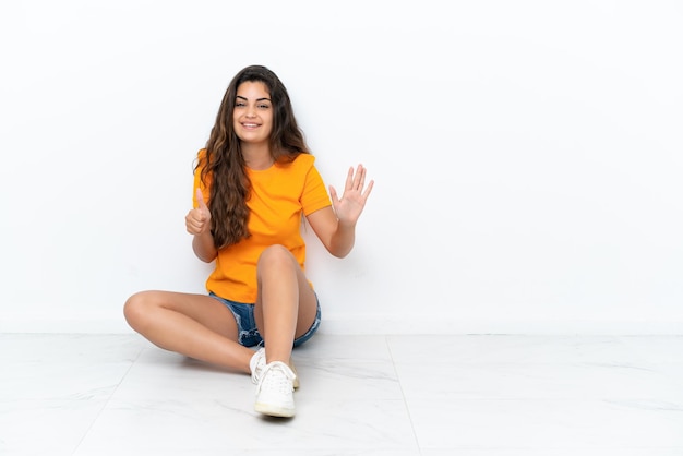 Foto joven mujer caucásica sentada en el suelo aislado sobre fondo blanco contando seis con los dedos