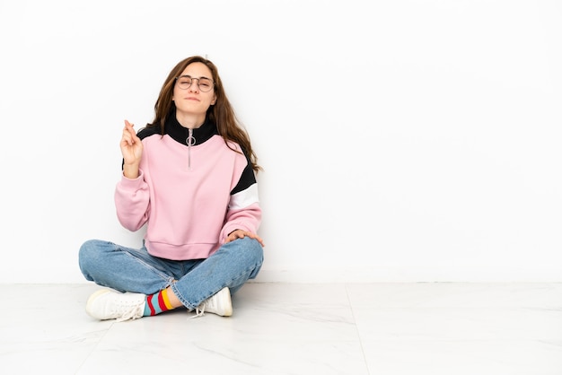 Joven mujer caucásica sentada en el suelo aislado sobre fondo blanco con los dedos cruzando y deseando lo mejor